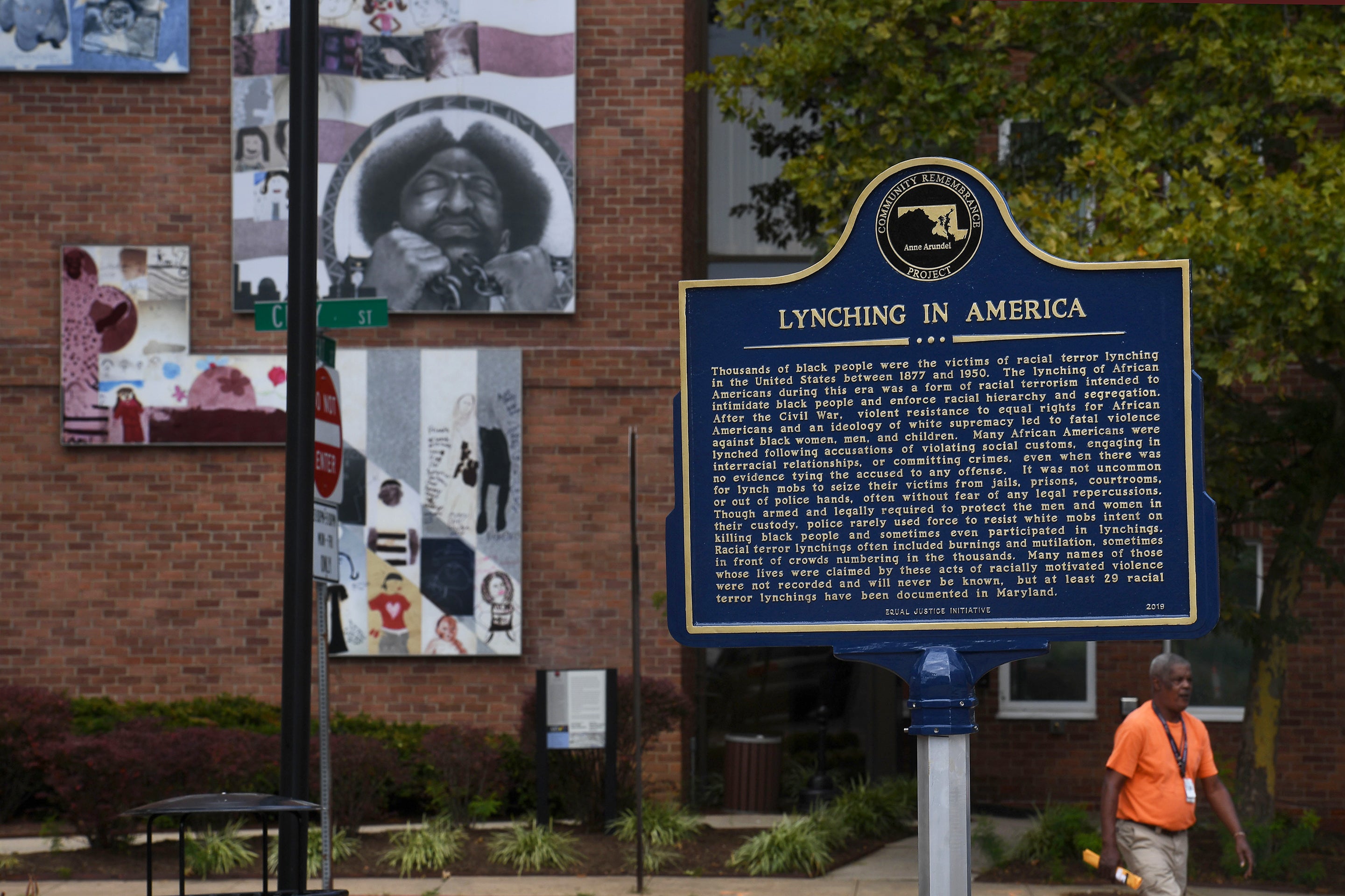 Un panneau historique détaillant le lynchage en Amérique est visible dans une rue d'Annapolis, Maryland, avec une œuvre d'art installée sur le mur d'un bâtiment en arrière-plan, alors qu'un homme non identifié passe sur le trottoir.