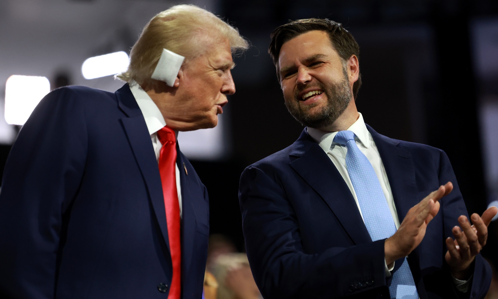 Donald Trump et J.D. Vance, en costume, se tiennent sur scène. Trump, avec un bandage blanc sur l'oreille, parle à Vance.