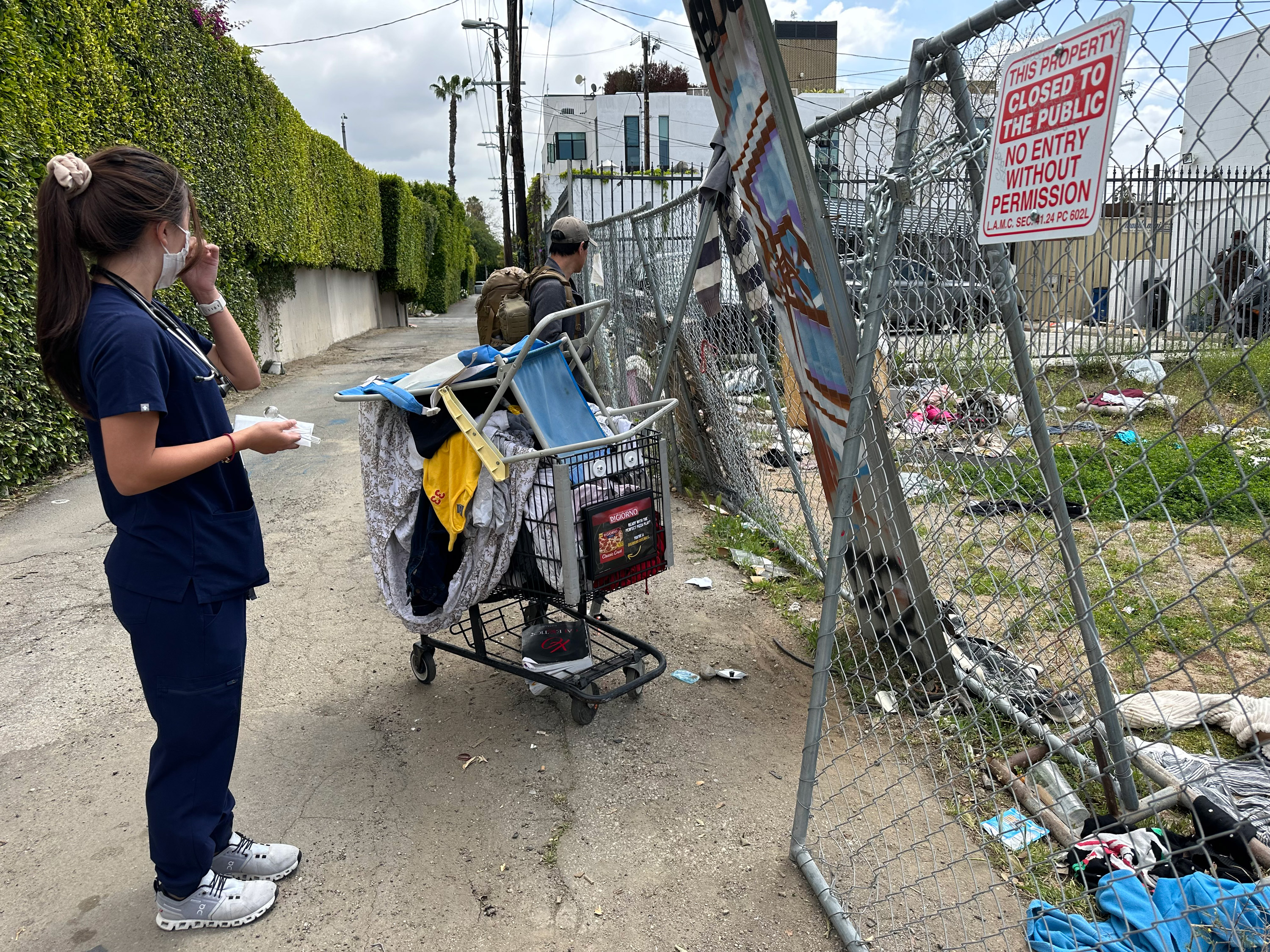 Une photo d'une médecin en uniforme regardant dans une ruelle. Elle est à côté d'un chariot de courses rempli d'affaires.