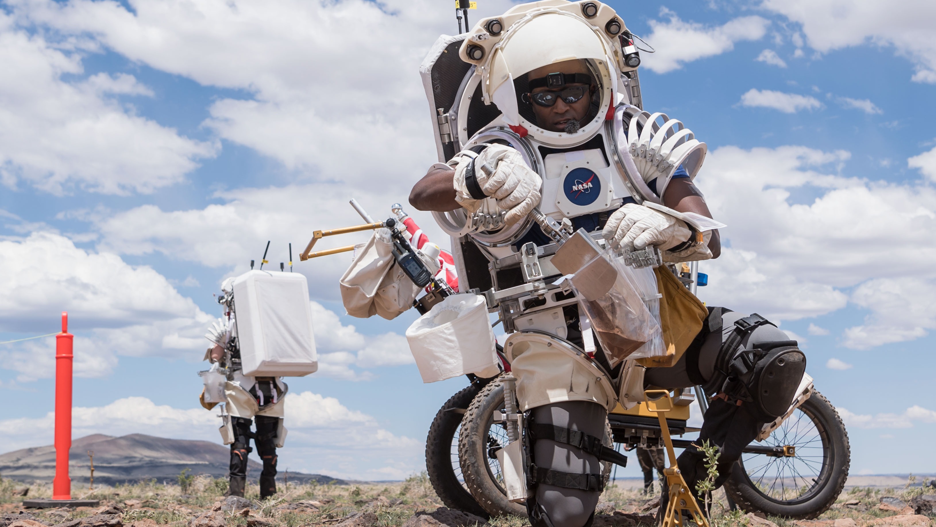 deux astronautes en tenue de simulation sur le terrain. L'un d'eux utilise un rover lunaire et des outils pour prélever un échantillon de sol. Des nuages et un ciel bleu sont visibles en arrière-plan