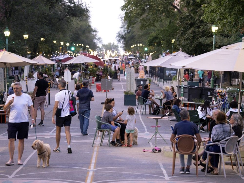 actualités Des gens se promènent dans une rue piétonne en été. Certains sont assis à des tables sous des parasols.