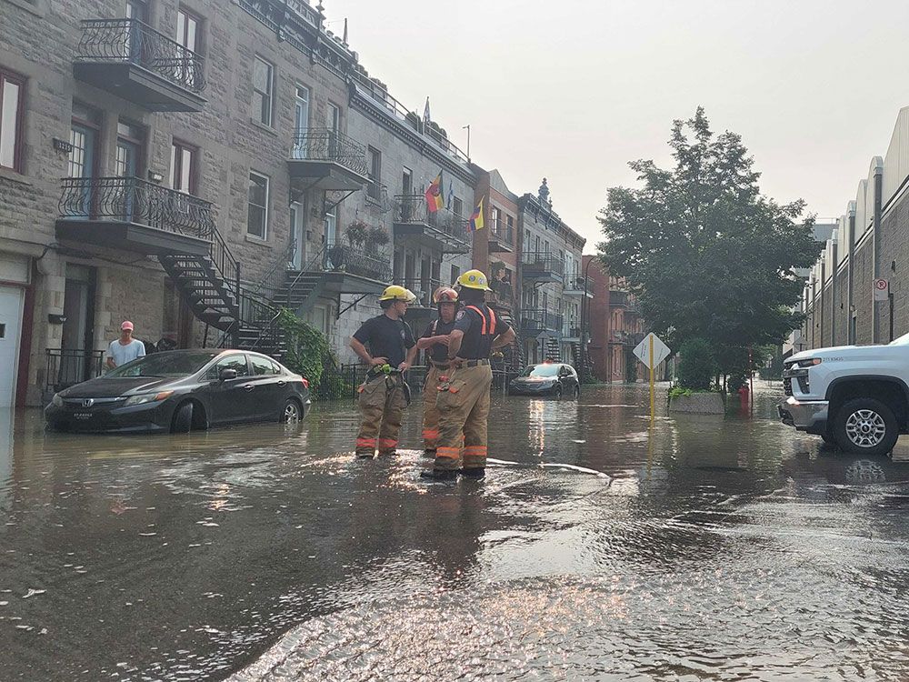 Trois pompiers se tiennent au milieu d'une rue inondée. L'eau leur arrive aux chevilles.