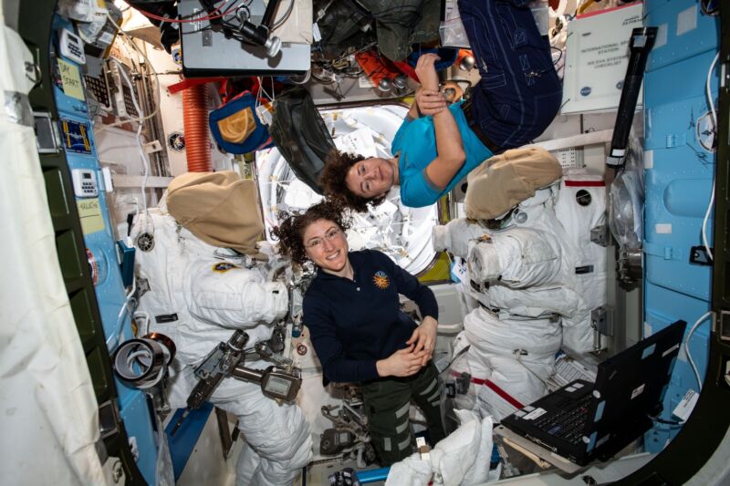 Image de deux astronautes dans une chambre remplie d'équipements, se tenant près des combinaisons qu'ils portent pour les activités extravéhiculaires.