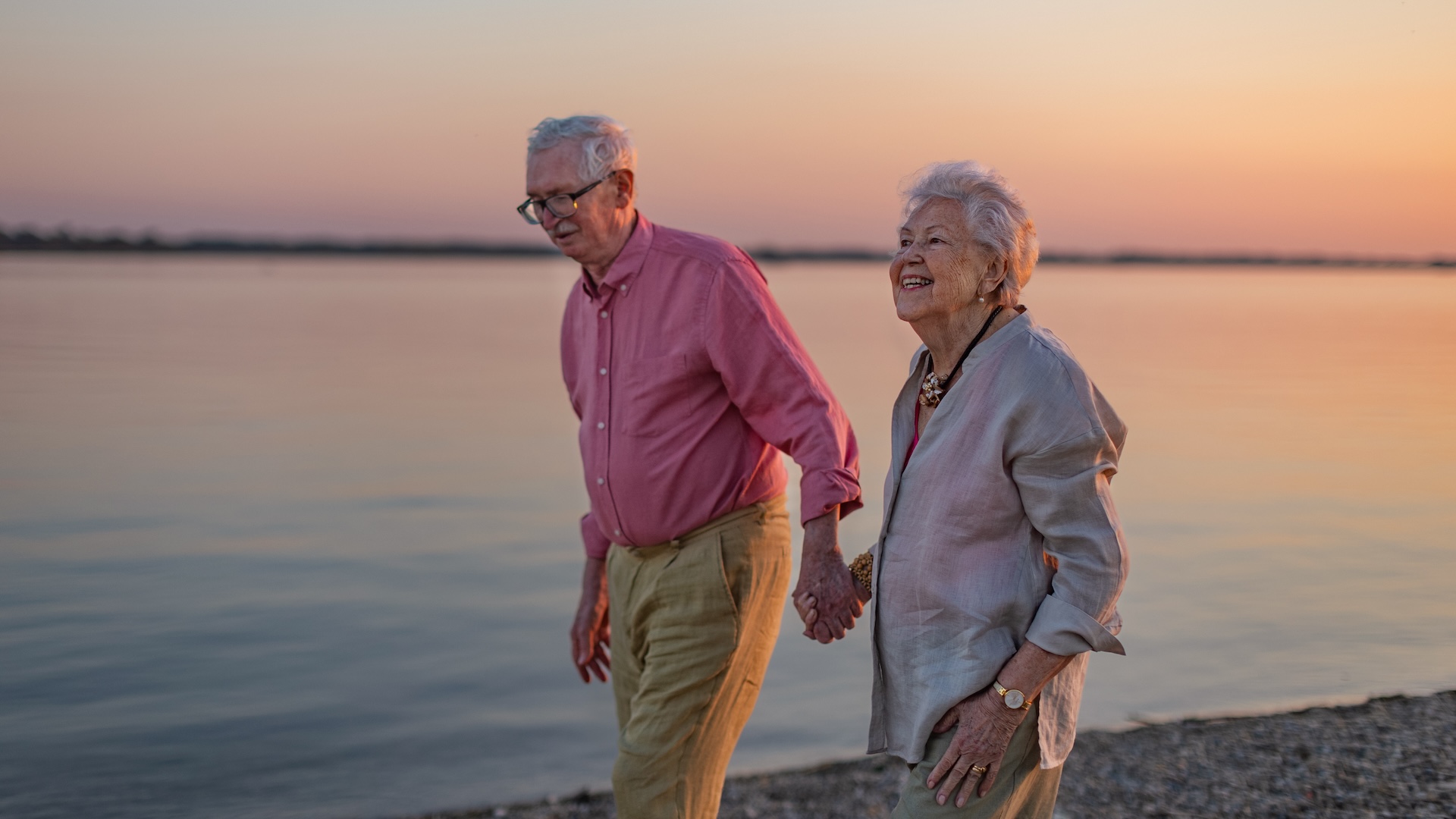 science Un couple âgé sourit en se promenant sur la plage main dans la main au coucher du soleil