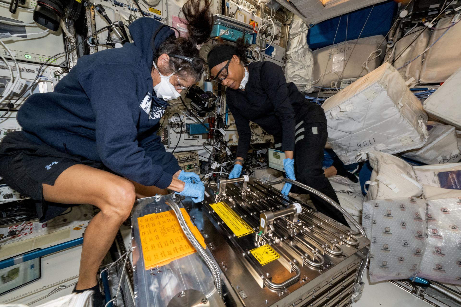 Deux femmes en survêtements déballent une grande boîte métallique dans un laboratoire exigu rempli de fils et d'ordinateurs. Elles flottent.