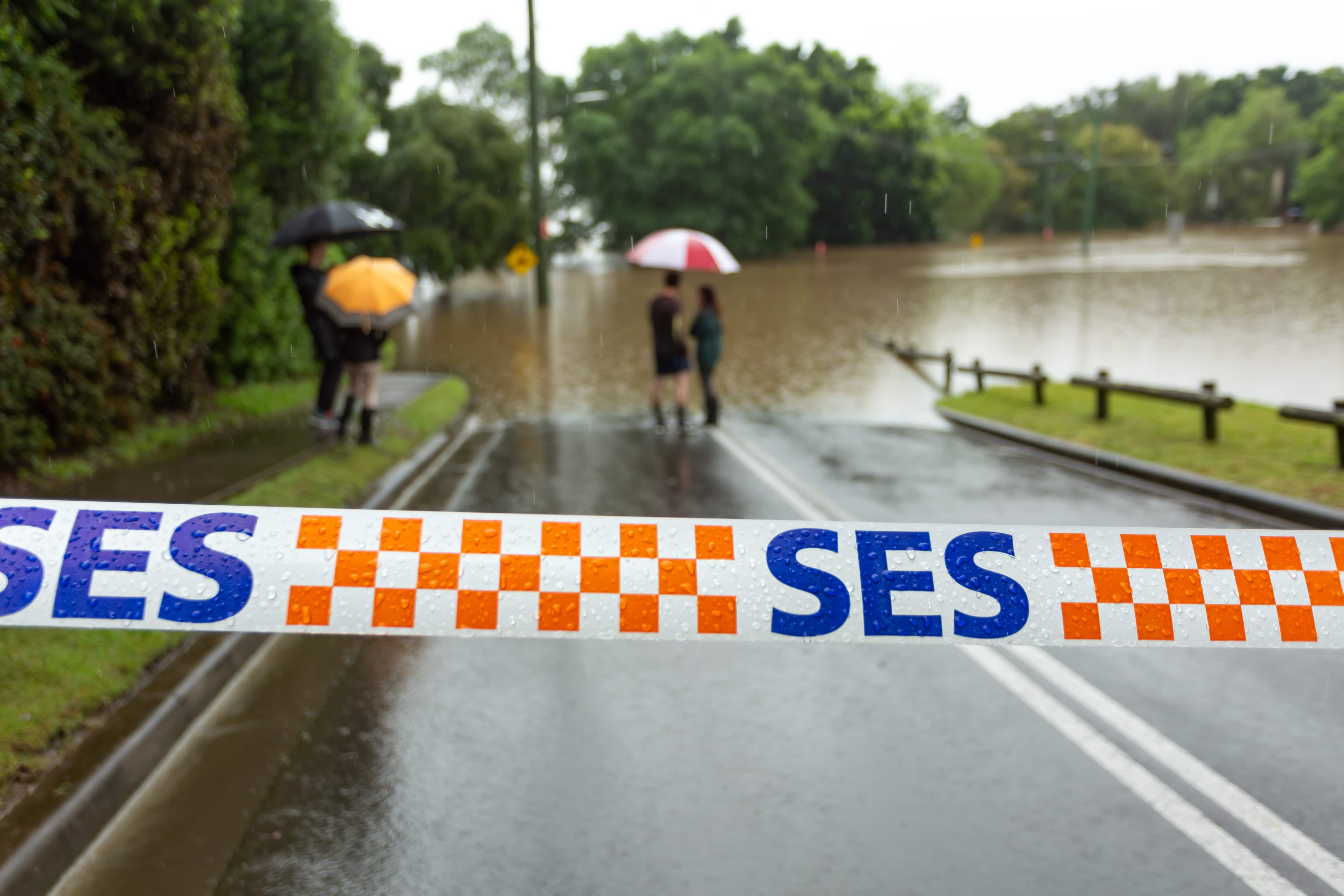 personnel des services d'urgence bloquant une route, où des personnes se tiennent près d'un site d'inondation avec des parapluies.